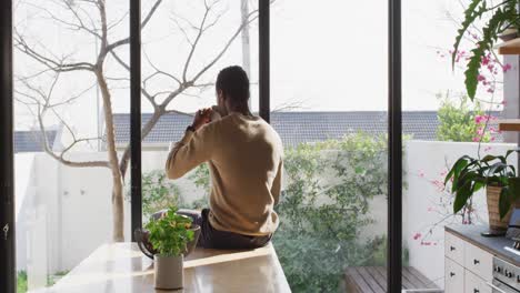 happy african american man sitting at countertop in kitchen, drinking coffee