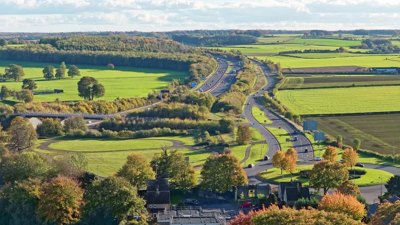 Wide aerial view of the A1(M) motorway through English countryside near Wetherby, Yorkshire. Multiple lanes, flowing traffic, open rural scenery, autumn foliage, peaceful panoramic