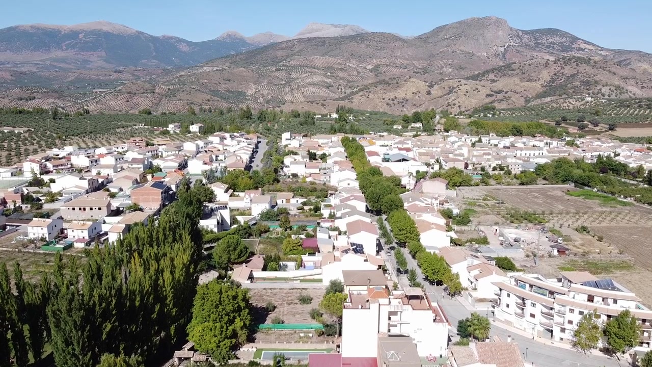 Immagini aeree registrate con un drone che mostrano Arbuniel, un tranquillo villaggio a Jaén, Andalusia, integrato in un paesaggio di uliveti e montagne aspre sotto un cielo soleggiato e luminoso.
