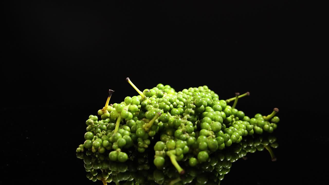 A cluster of fresh green peppercorns rotates smoothly on a reflective black surface, illuminated by dramatic studio lighting with a seamless, high-contrast look