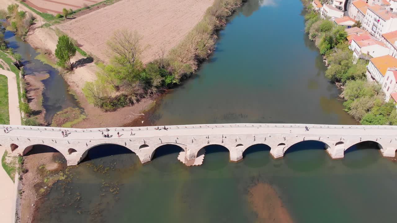 Aerial view of an ancient stone arch bridge spanning a river, with people walking and cycling