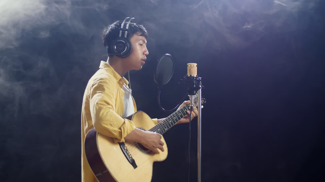 하 연기 검은 배경에 기타를 연주하고 콘덴서 마이크로 노래하는 헤드폰을 가진 젊은 아시아 소년의 측면 시각