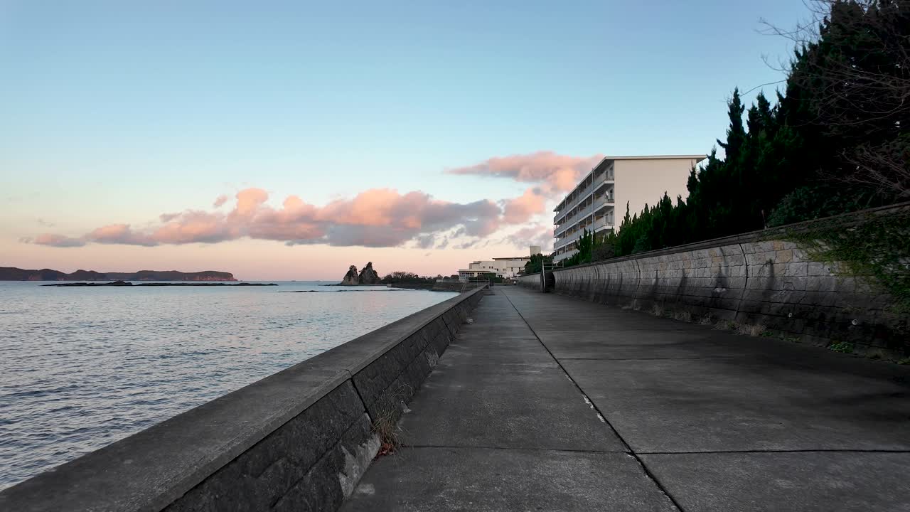 First person view walking on a coastal path beside Nachi Bay during golden hour, with pink clouds and calm sea
