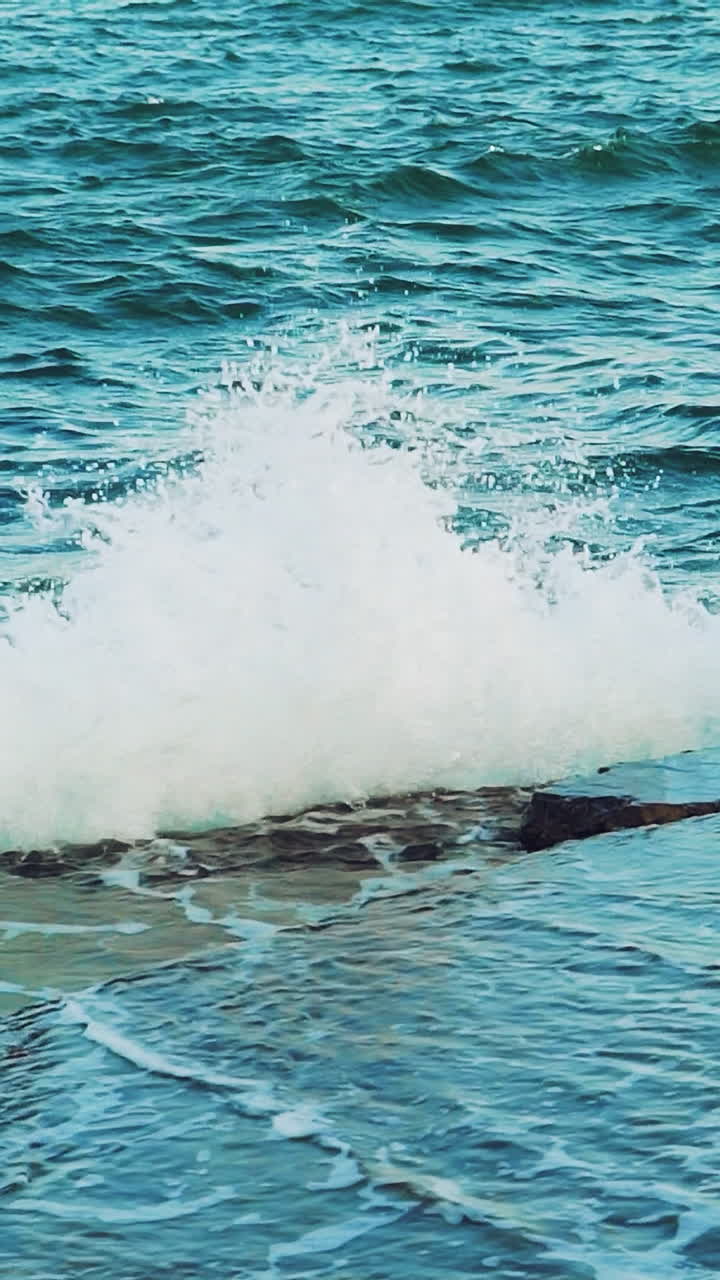 Turquoise waves is colliding with stone rocks near the shore and turning into thousands of drops of water. Slow motion. View from above Vertical video