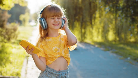 Happy-child-girl-in-wireless-headphones-choosing,-listening-music-in-smartphone-dancing-outdoors