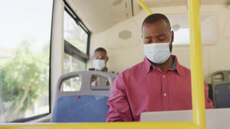 hombre afroamericano en la ciudad con máscara facial, usando computadora portátil en el autobús