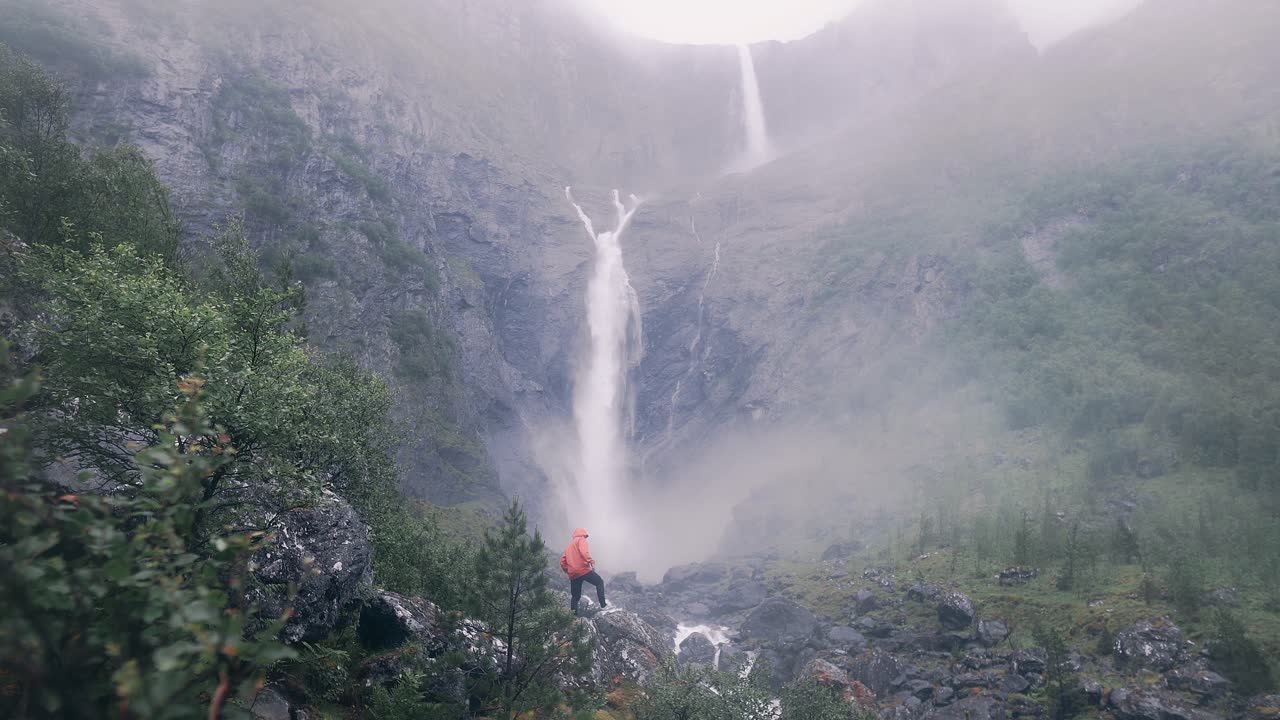 Young man standing in front of a large waterfall, wearing a red jacket, steam around from the water. Surrounded by mountains.