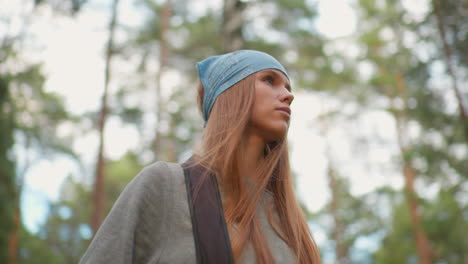 young woman hiking in lush forest, gazing thoughtfully downward, wearing blue bandana and holding backpack straps, surrounded by vibrant greenery and tall trees