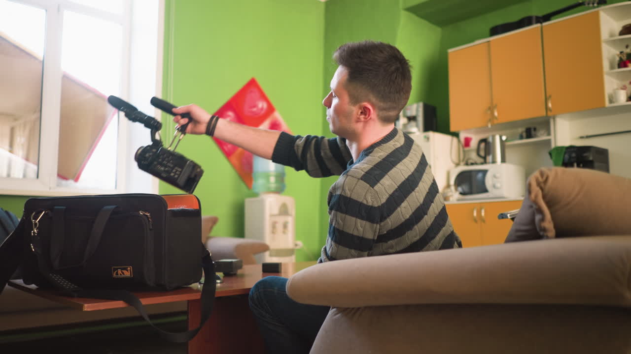 Uomo che cerca un microfono in una borsa nera per attrezzatura fotografica mentre è seduto a un tavolo in un ufficio domestico con pareti verdi. Attrezzatura fotografica, obiettivi e altre apparecchiature visibili sul tavolo nell'area di lavoro.