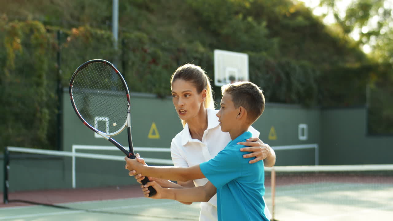 Smiling Woman Teaching Her Son How To Hit Ball With Racket On A Tennis Court