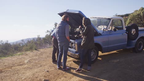 A-group-of-friends-stand-around-an-old-blue-pickup-truck-with-its-hood-up-on-the-side-of-a-rural-road