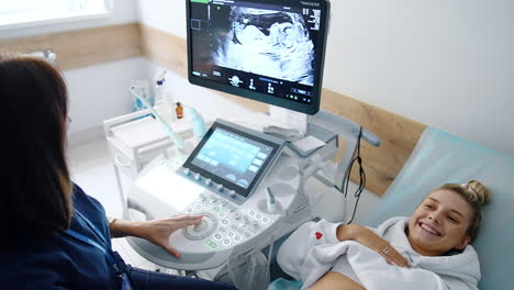 happy excited lady lies on the couch near ultrasound equipment. beautiful pregnant woman points at the screen seeing her baby.