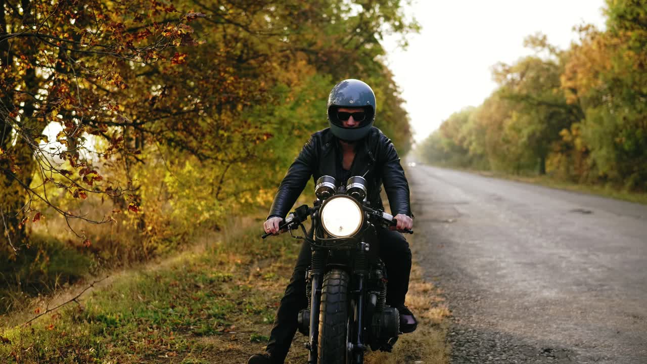 hombre irreconocible con casco negro y chaqueta de cuero deteniendo su motocicleta vintage en el lado de la carretera en un soleado día de otoño
