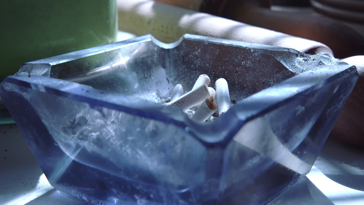 Close up of a blue glass ashtray with a person's hand tapping the ash off a cigarette