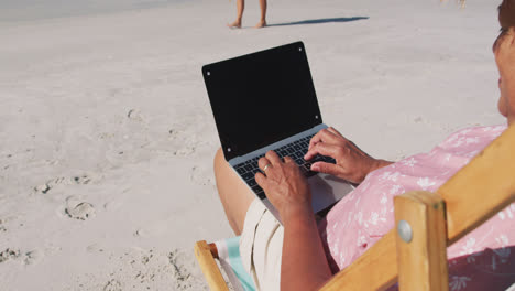 Mujer-Mayor-De-Raza-Mixta-Sentada-En-Una-Tumbona-Y-Usando-Una-Computadora-Portátil-En-La-Playa