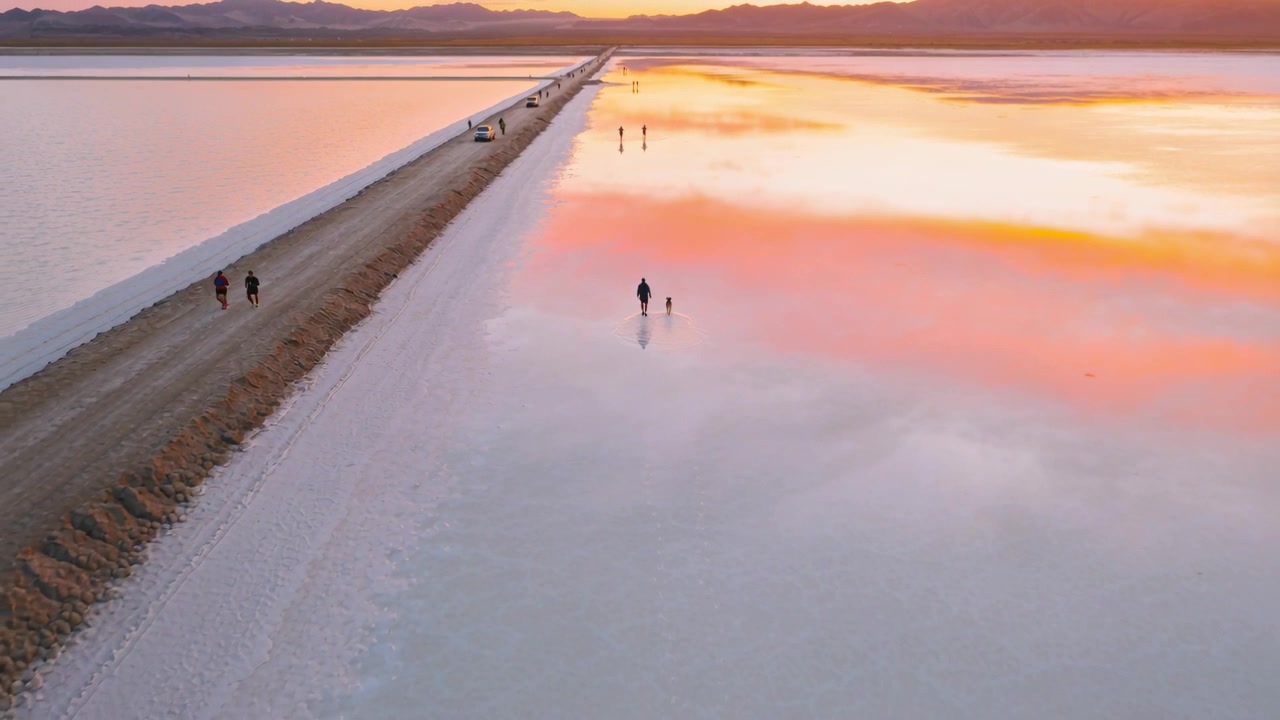 Recording walker in dark coat and dog crossing salt flat beside causeway at sunset, reflecting sky