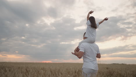 daddy carries on his shoulders his beloved little healthy daughter in sun. in slow motion the daughter walks with her father on the field and free and happy waves her hands up. walking in field.