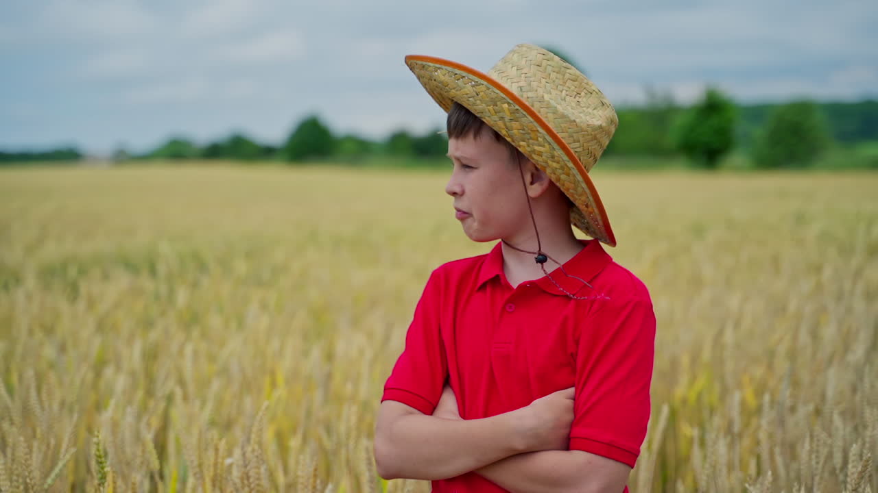 Teenager in straw hat among nature. Portrait of a pensive boy standing on a wheat field in summer. Serious child on the background of yellow farmland.