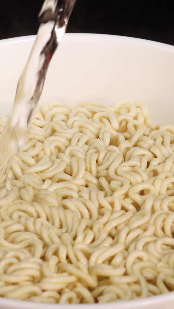 白い器に入ったインスタントラーメンに、均一でニュートラルな照明の下、熱いお湯が絶え間なく注がれる様子をクローズアップで捉えています。