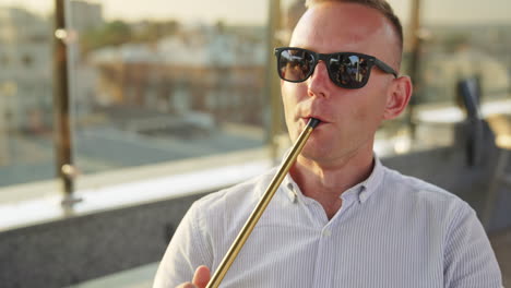 mid aged man in sunglasses holding shisha pipe in his mouth. close up. relaxed man in shirt smoking hookah in the café outdoors.