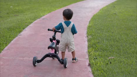 Junger-Lateinamerikanischer-Junge-Mit-Overalls,-Der-An-Einem-Warmen-Nachmittag-Sein-Fahrrad-Auf-Der-Straße-Im-Park-Schleppt