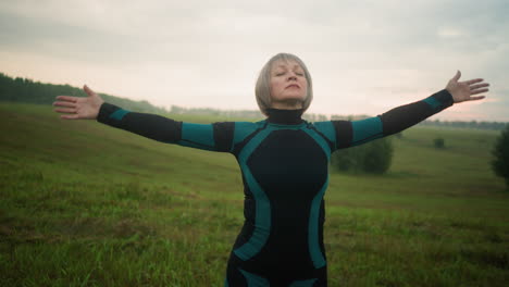 woman in green black suit stands with head slightly lifted, eyes closed, and hands slowly outstretched and brought down in a serene motion, practicing yoga in a vast open field under a cloudy sky