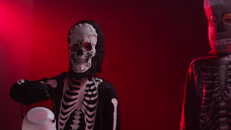 fotografía de estudio de niños vestidos con trajes de esqueleto haciendo trucos o tratamientos con cubos de caramelos en halloween con iluminación de fondo de humo rojo 1