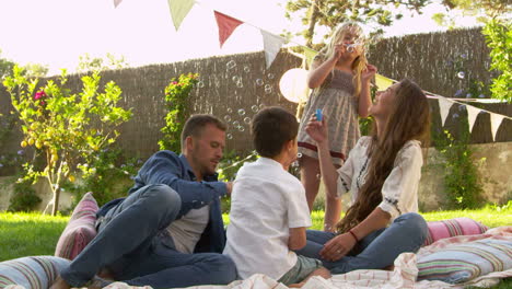 Family-Sitting-On-Blanket-In-Garden-Blowing-Bubbles