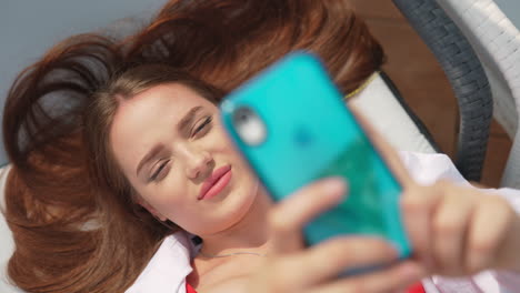 brunette girl sunbathing in a chaise lounge close up. beautiful lady holding phone over her face, taking selfie.