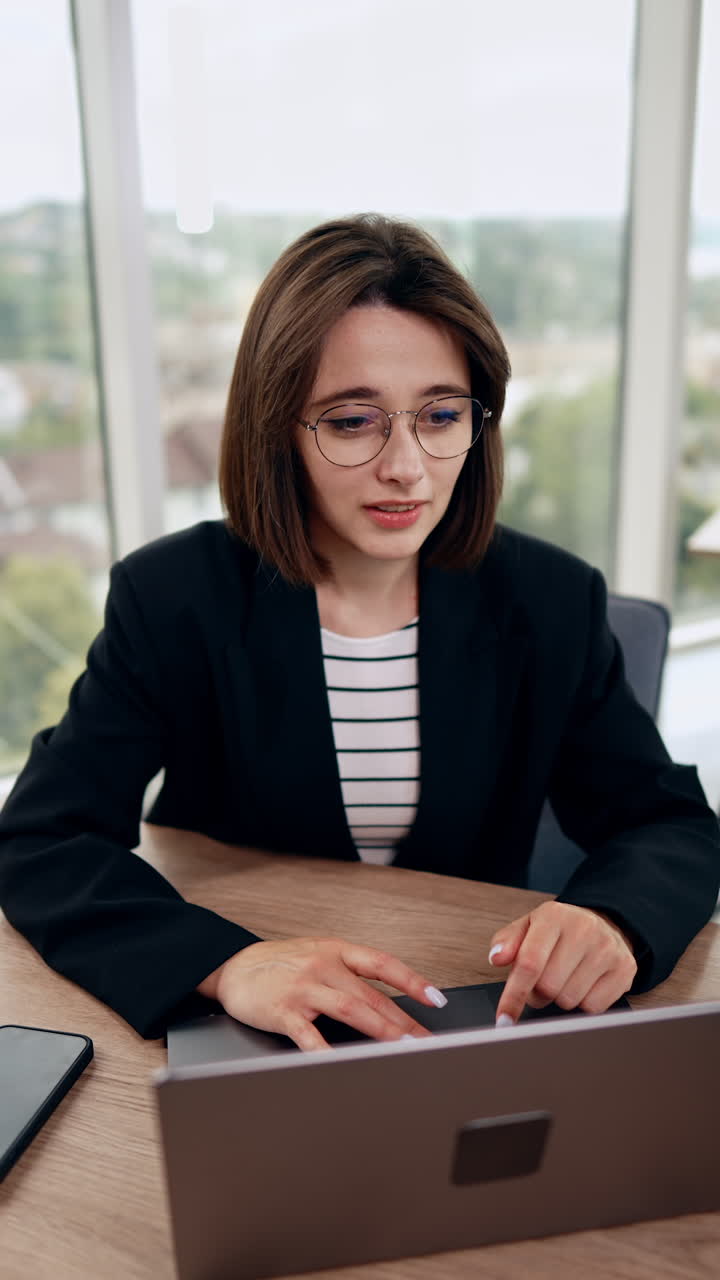 Smiling Caucasian lady in glasses works on laptop. Top view on the woman working in the office. Vertical video.
