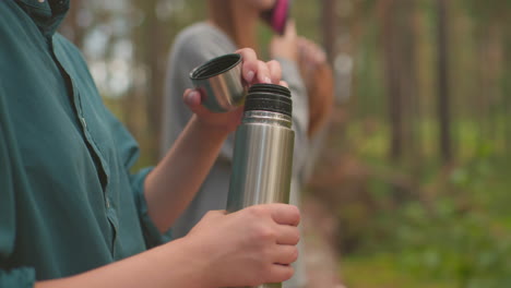 friends takes break in lush forest, one carefully opens thermos while other gently brushes hair, backpacks rest on fallen tree, capturing relaxed, serene moment as they enjoy nature