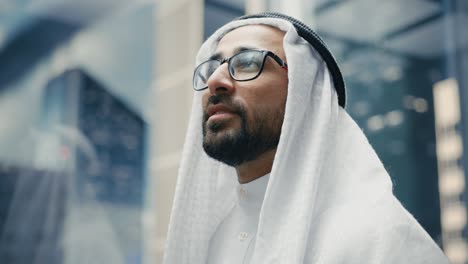 portrait of a successful happy muslim businessman in traditional white kandura wearing glasses riding glass elevator to office in modern business center. saudi, emirati, arab businessman concept.