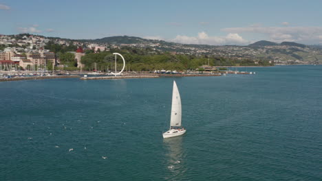 Filmische-Luftaufnahme-Eines-Bootes,-Das-über-Den-Genfersee-Segelt,-Mit-Einer-Wunderschönen-Skyline-Der-Stadt-Im-Hintergrund