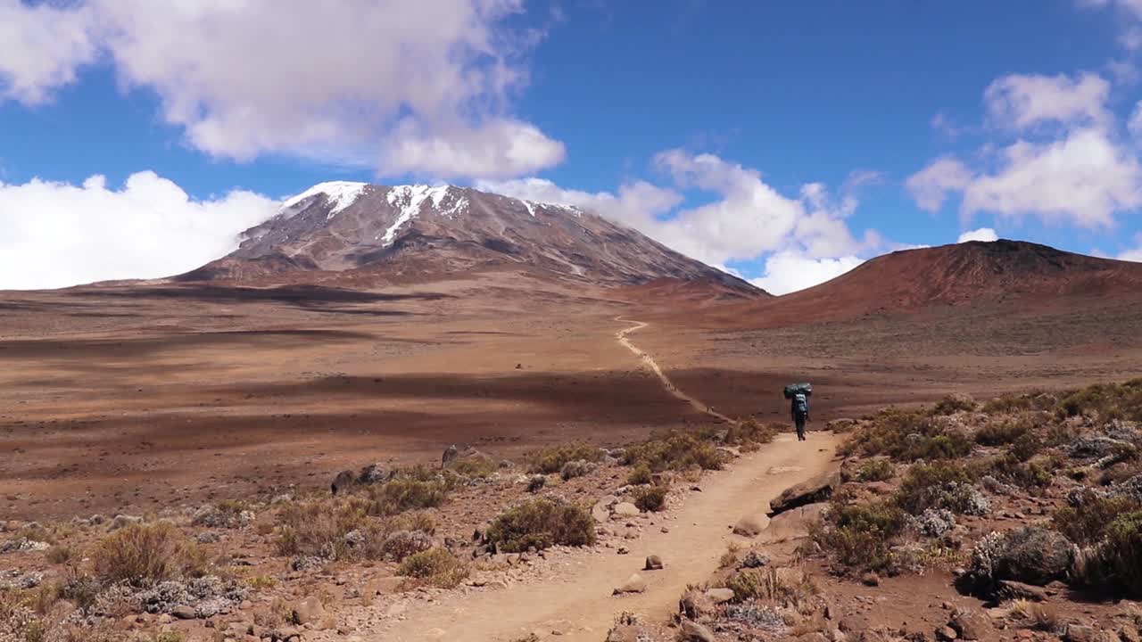 toma estática de un hombre caminando hacia la cima del monte kilimanjaro, cargando cosas en la cabeza, en un día soleado, cerca de la cabaña horombo, en tanzania, áfrica.