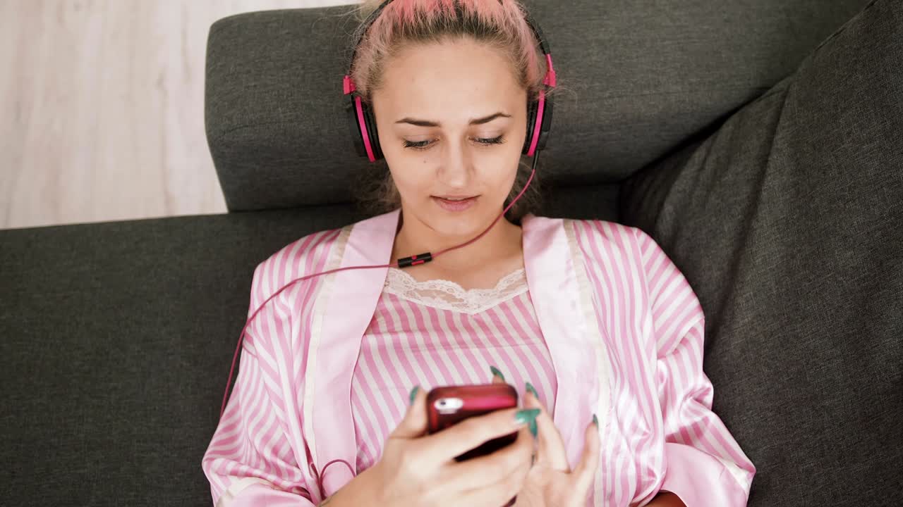 Top view portrait of an beautiful young woman with pink hair laying down on bed and listen to music with headphones