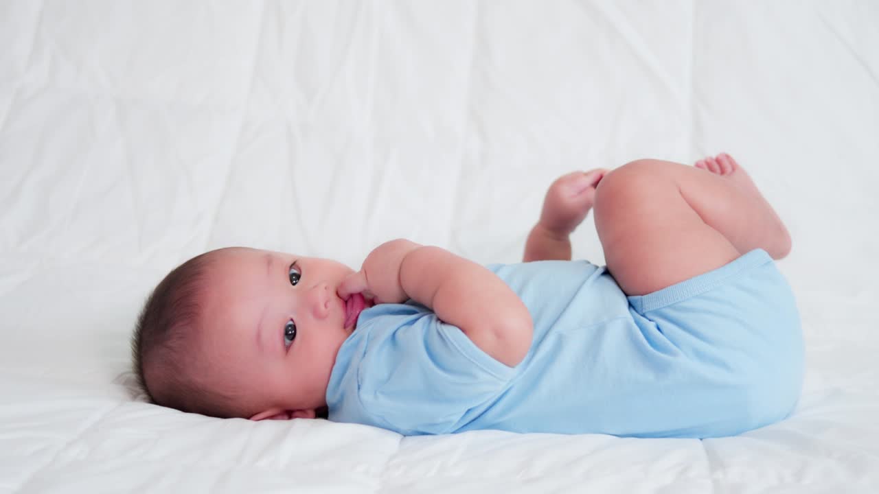 familia feliz, lindo bebé recién nacido asiático usar camisa azul acostado jugando en la cama blanca mirando a la cámara