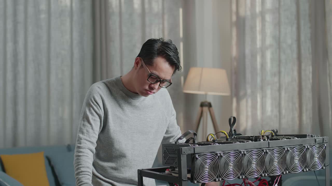 안경을 쓴 아시아인 남자가 머리를 흔들고 노트북과 광산 기계를 사용하여 암호화폐를 채굴하는 동안 두통을 고 있습니다.