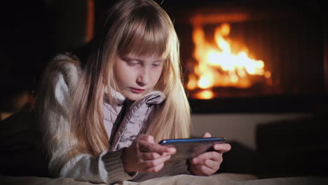 Blonde-Girl-Uses-A-Smartphone-Lies-On-The-Floor-In-The-Room-Against-The-Background-Of-The-Fireplace