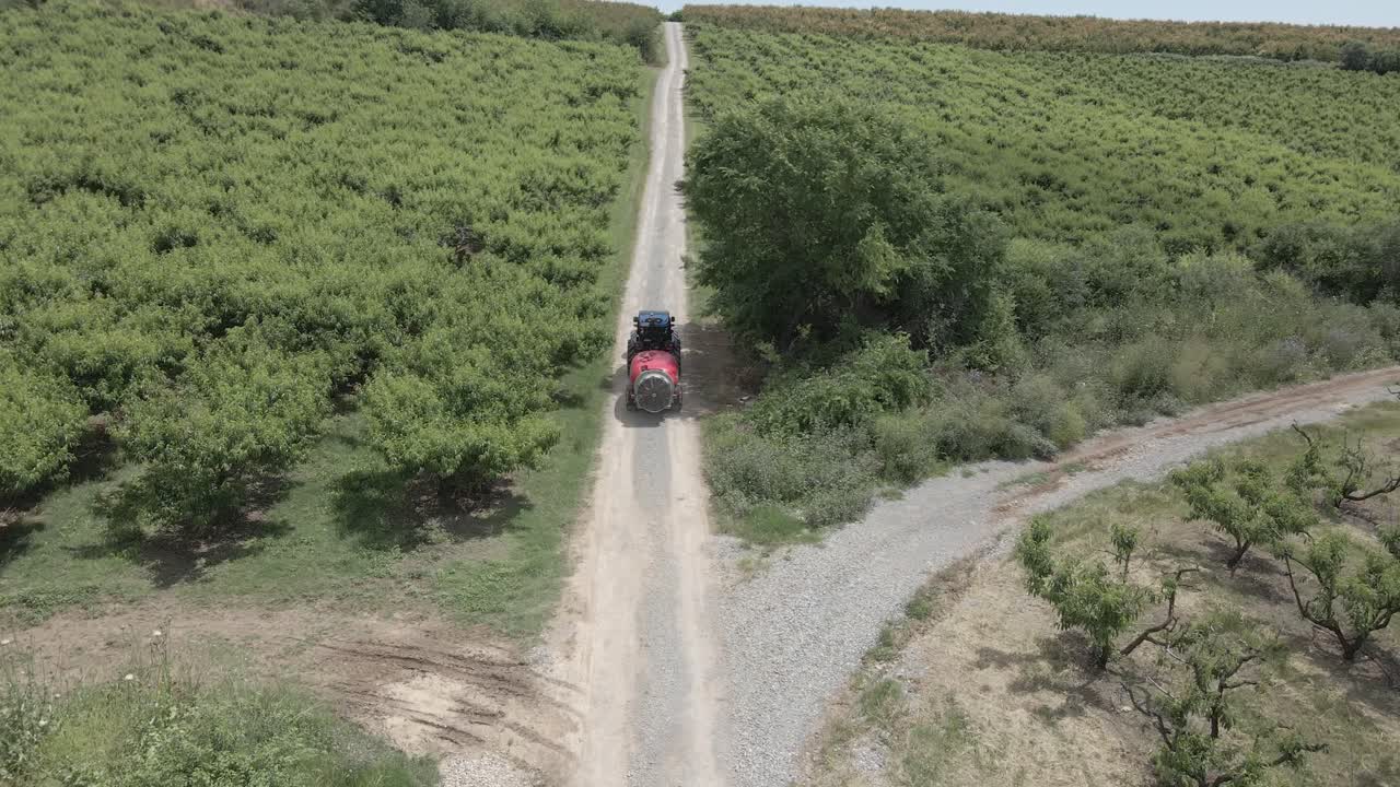 Aerial 4K footage at 50fps of a blue tractor pulling a red sprayer tank along a rural dirt road in a lush orchard, showcasing modern agricultural machinery in a scenic hilly landscape.