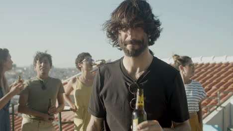 retrato de un hombre caucásico barbudo mirando la cámara y bebiendo cerveza mientras tiene una fiesta en la azotea con amigos