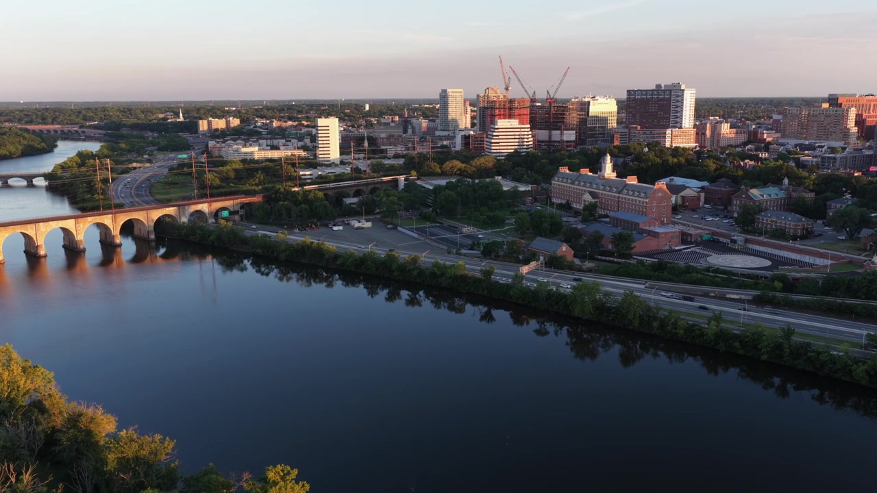 Un ponte ad arco in pietra attraversa un fiume calmo verso New Brunswick, New Jersey al tramonto, gru edili svettano sopra lo skyline del centro accanto a storici edifici universitari in mattoni. Ampia ripresa aerea con drone