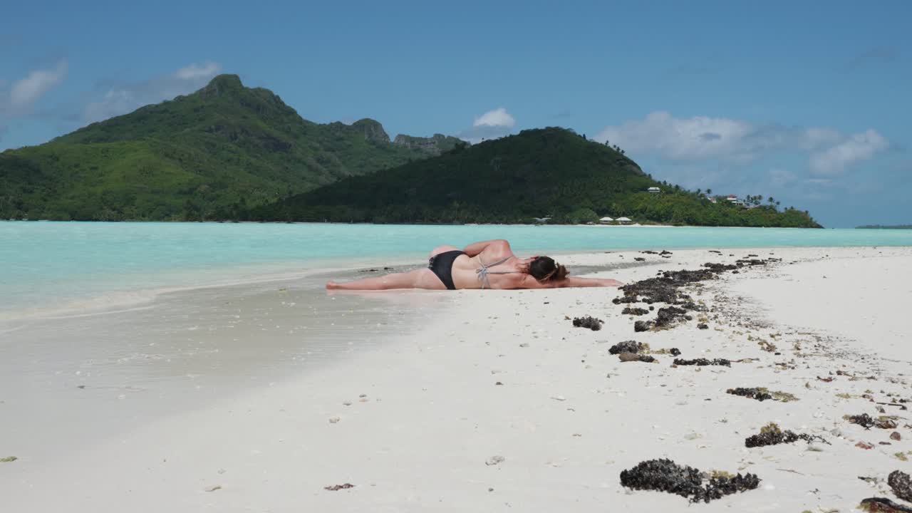Young caucasian woman in bikini and sunglasses laying down, sunbathing and relaxing on a white sand tropical beach with feet in the water and green pacific island behind in Maupiti, French Polynesia.