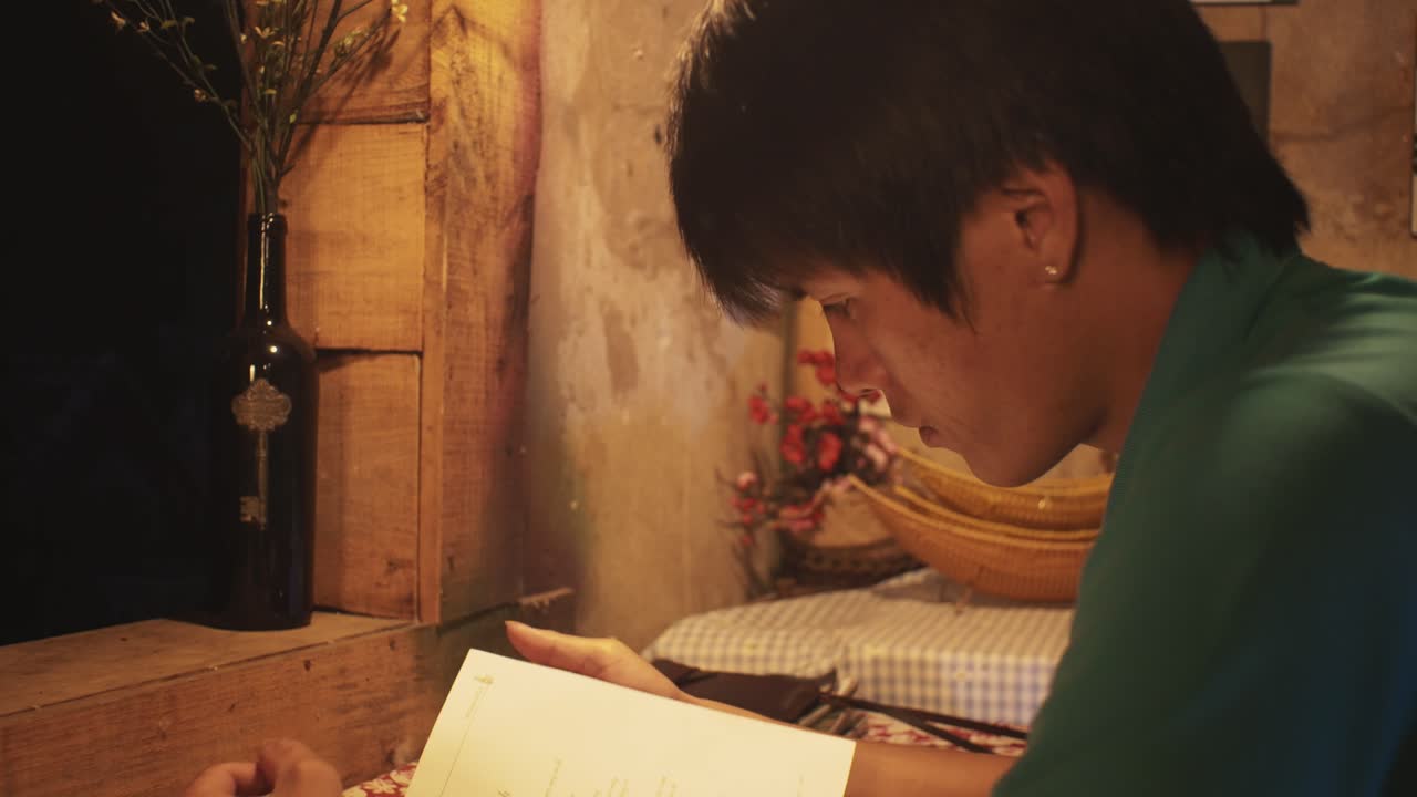 Young Asian male reads a book at a desk in a rustic room
