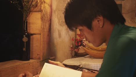 young asian male reads a book at a desk in a rustic room