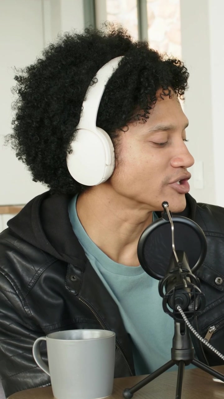 Video verticale: Conduttore in giacca che indossa cuffie bianche, si china verso il microfono in studio, registrando un podcast.