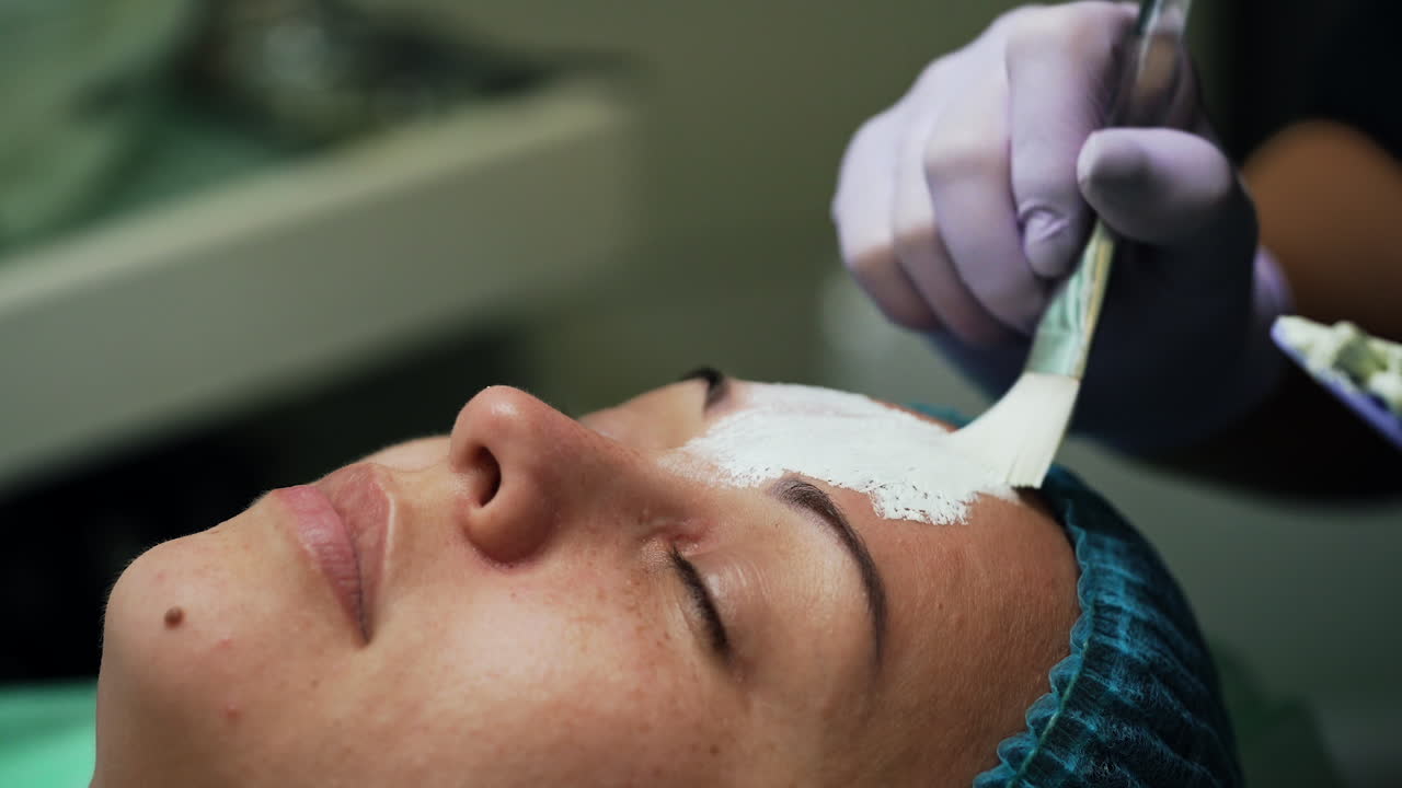 Beautician in latex gloves uses brush to cover client's face with mask. Rejuvenation procedures in beauty salon. Close up.