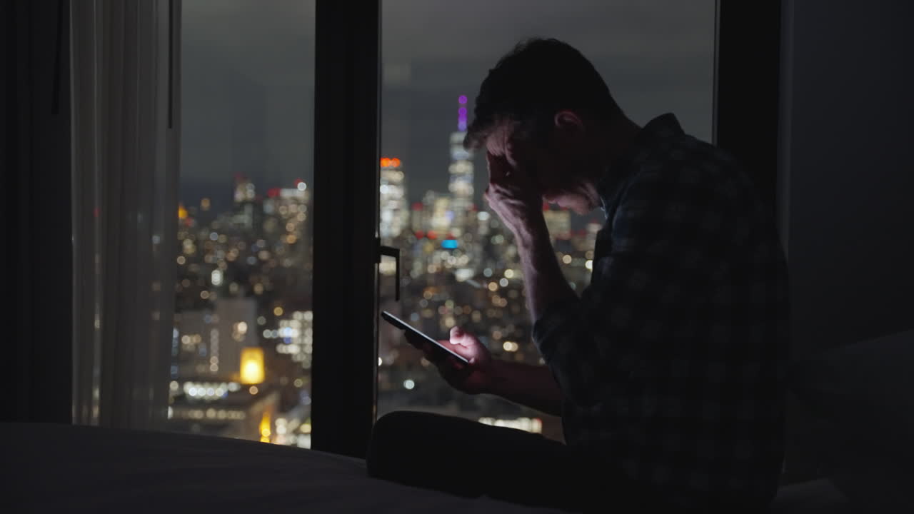 Uomo seduto sul letto che tiene in mano il telefono cellulare di notte, deluso e che fa gesti di incredulità dopo aver letto un messaggio o guardato un video