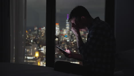 Man-sitting-on-bed-holding-mobile-phone-at-night-disappointed-and-making-disbelief-gestures-after-reading-a-message-or-watching-video