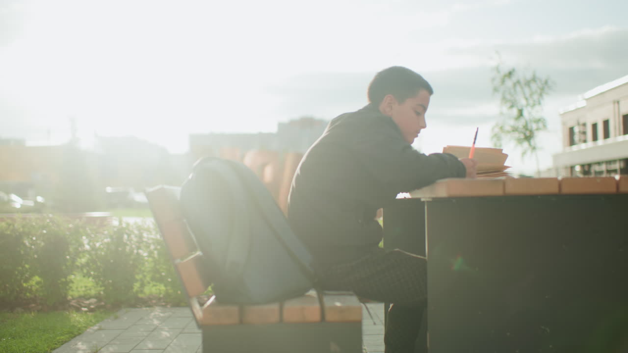 Side view of student writing with pen on notebook while sitting outdoors at wooden bench table near urban school building, backpack placed beside