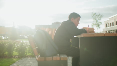 side view of student writing with pen on notebook while sitting outdoors at wooden bench table near urban school building, backpack placed beside